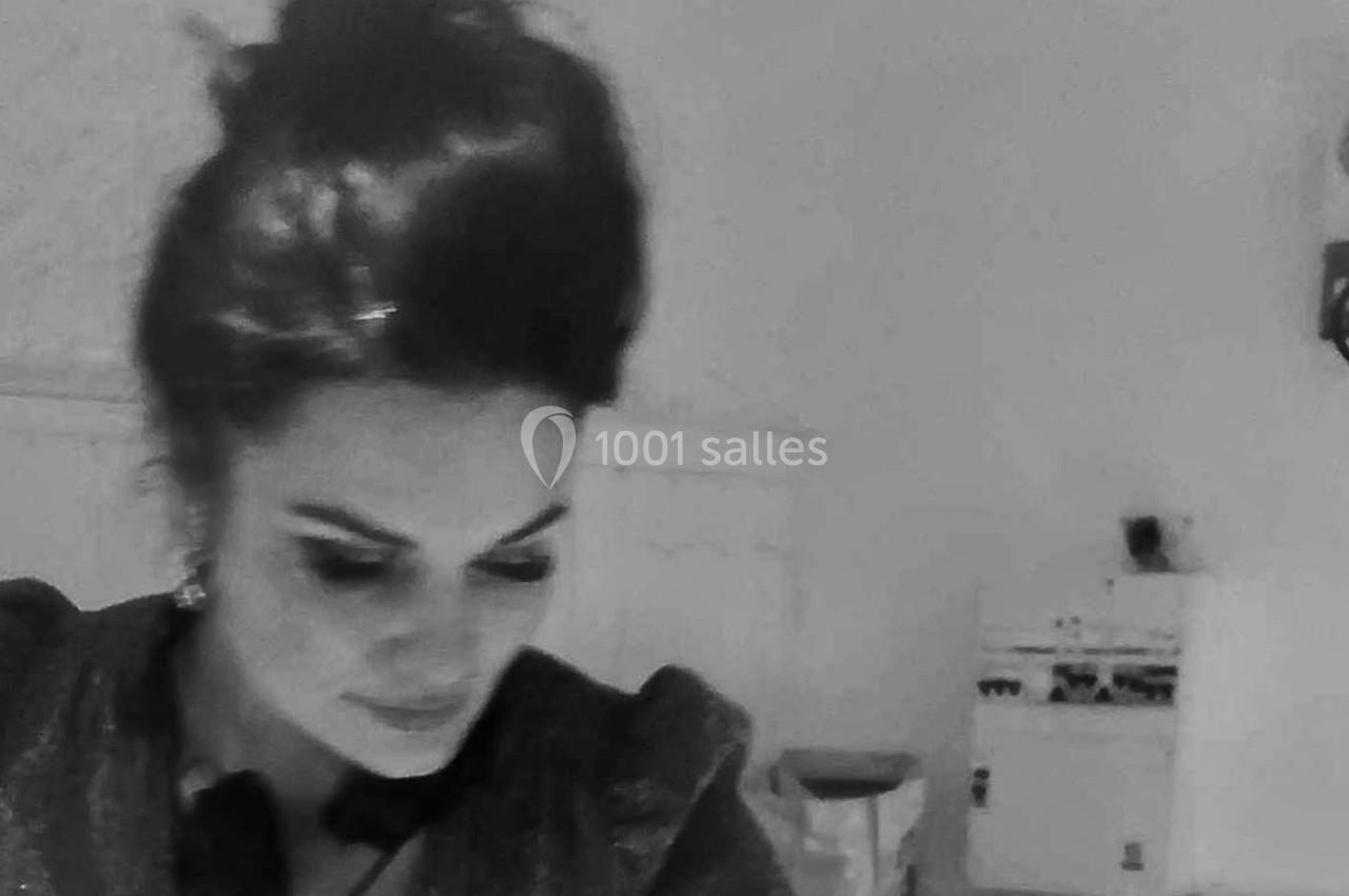 Femme aux cheveux relevés, regard baissé, dans une pièce sobrement éclairée avec des meubles en arrière-plan.