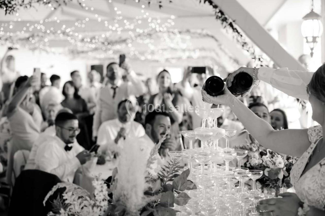 Des invités applaudissent tandis qu'une mariée remplit une pyramide de coupes avec du champagne lors d'une réception.
