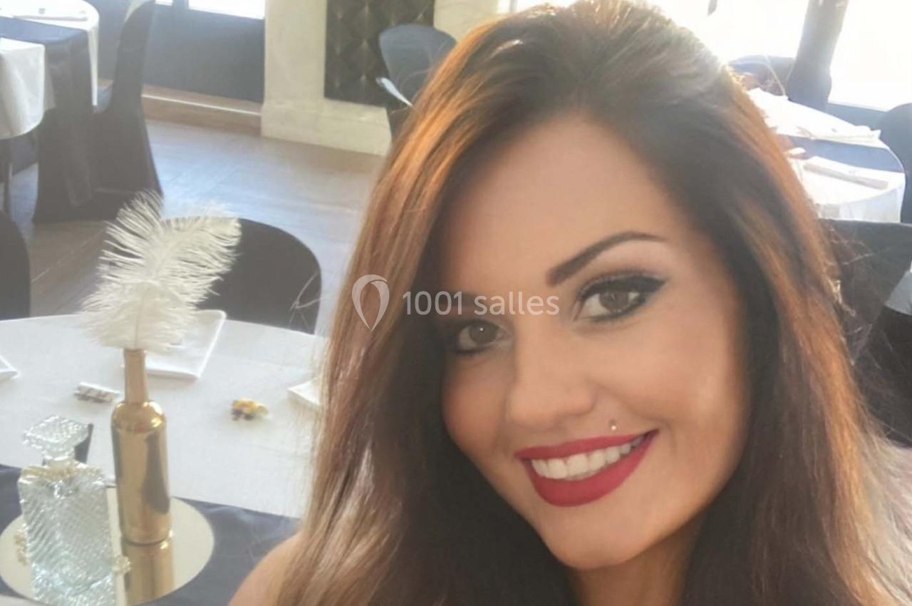 Une femme souriante avec des cheveux bruns, maquillage soigné, assise dans une salle décorée avec des tables dressées.