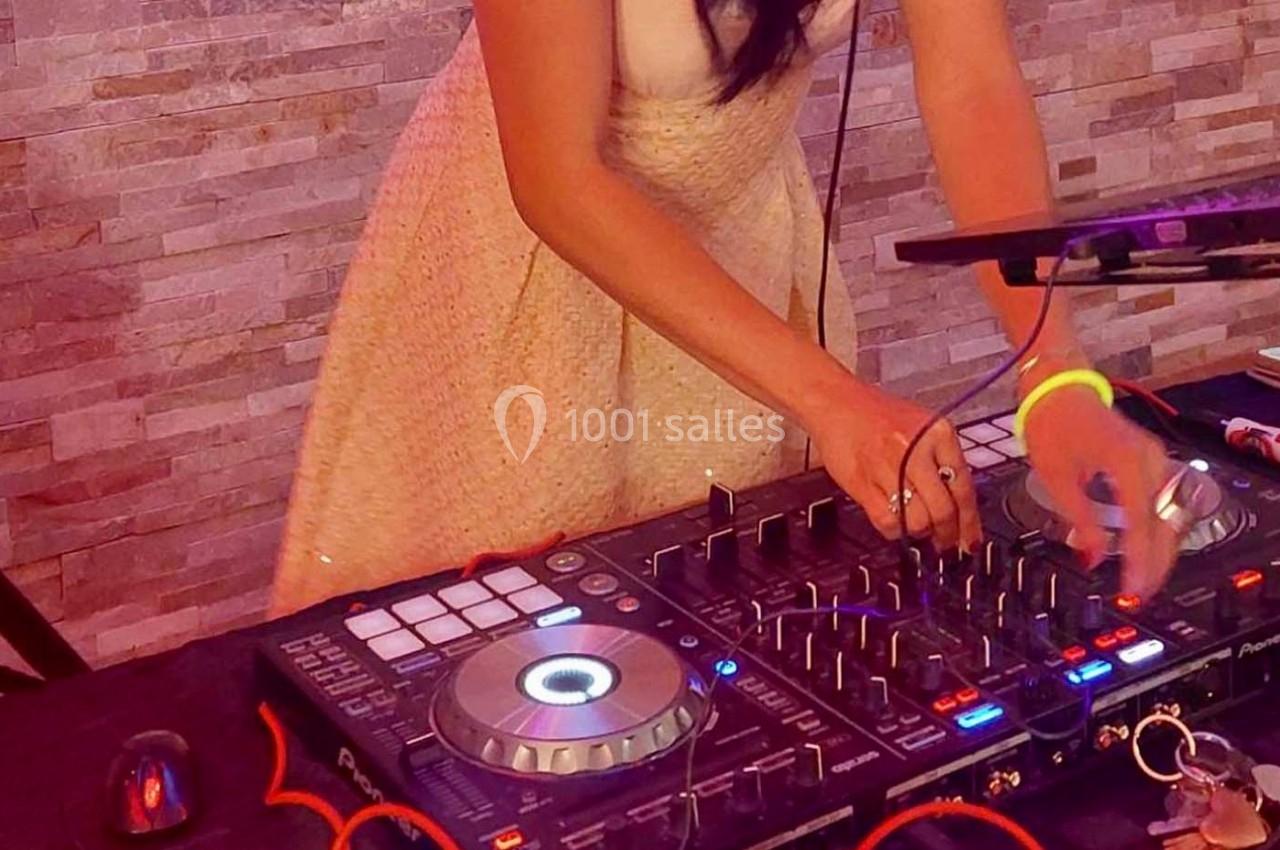 Une femme en robe claire mixe de la musique sur une table de mixage éclairée, devant un mur en pierre.