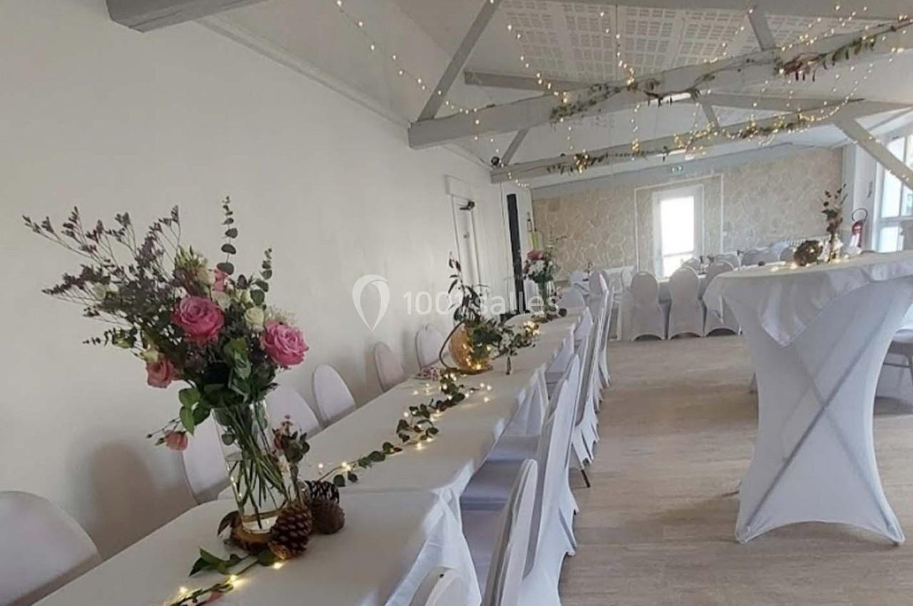 Salle décorée pour un événement avec tables alignées, nappes blanches, guirlandes lumineuses et bouquets de fleurs.