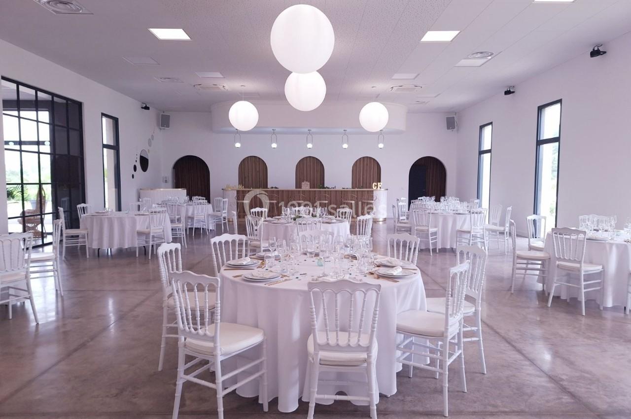 Salle de réception lumineuse avec tables rondes dressées, chaises blanches et suspensions sphériques au plafond.