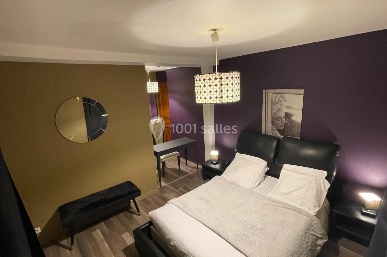 Chambre moderne avec lit double, murs violets et verts, miroir rond, bureau et éclairage suspendu.