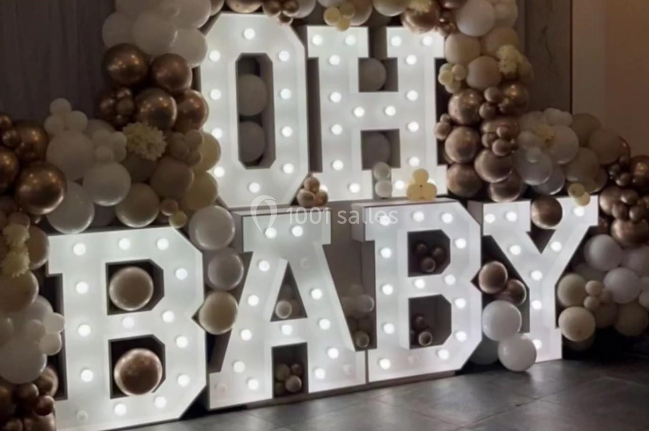 Lettres lumineuses ’OH BABY’ entourées de ballons blancs, dorés et beiges dans une décoration festive.