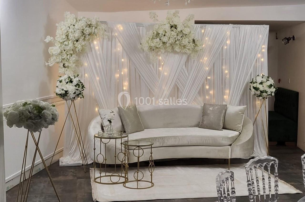 Canapé beige devant un fond drapé blanc décoré de fleurs blanches et de guirlandes lumineuses, avec tables dorées.