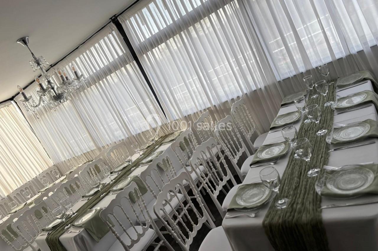 Salle de réception avec tables dressées, nappes blanches, chemins de table verts et chaises blanches, éclairée par un lustre.