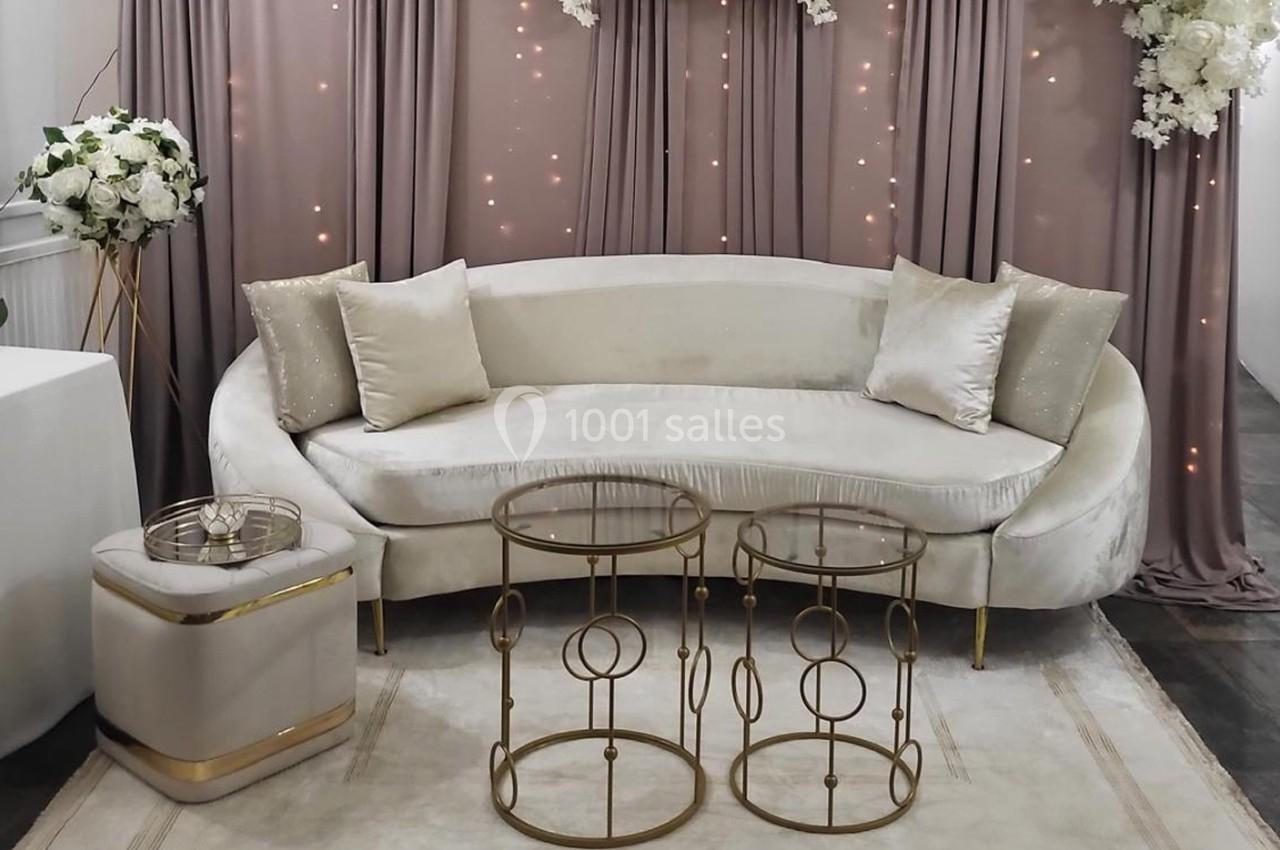 Canapé beige avec coussins, tables basses dorées et rideaux mauves décorés de lumières et fleurs blanches en arrière-plan.