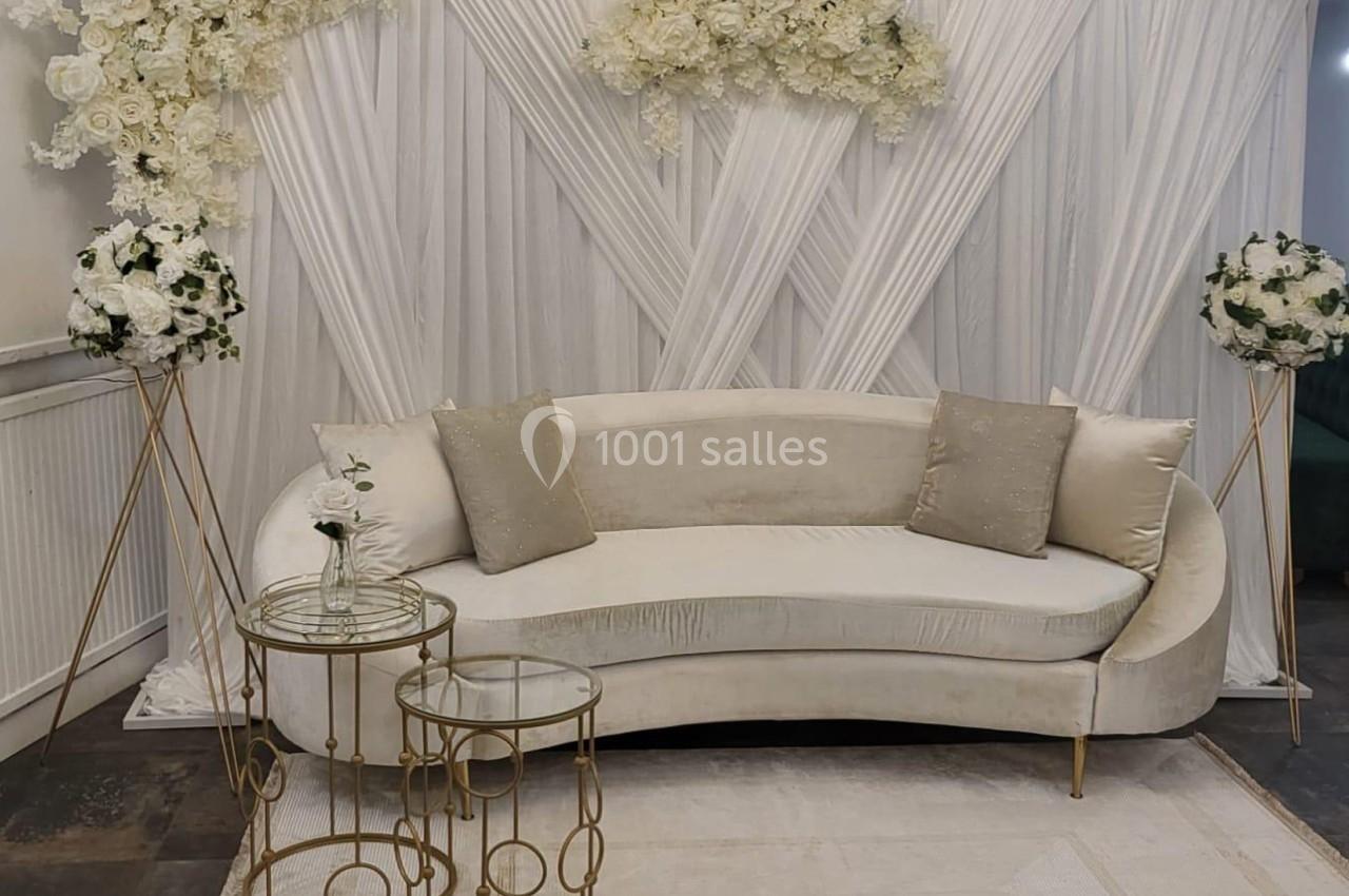 Canapé beige incurvé avec coussins assortis, entouré de fleurs blanches et de rideaux plissés en arrière-plan.