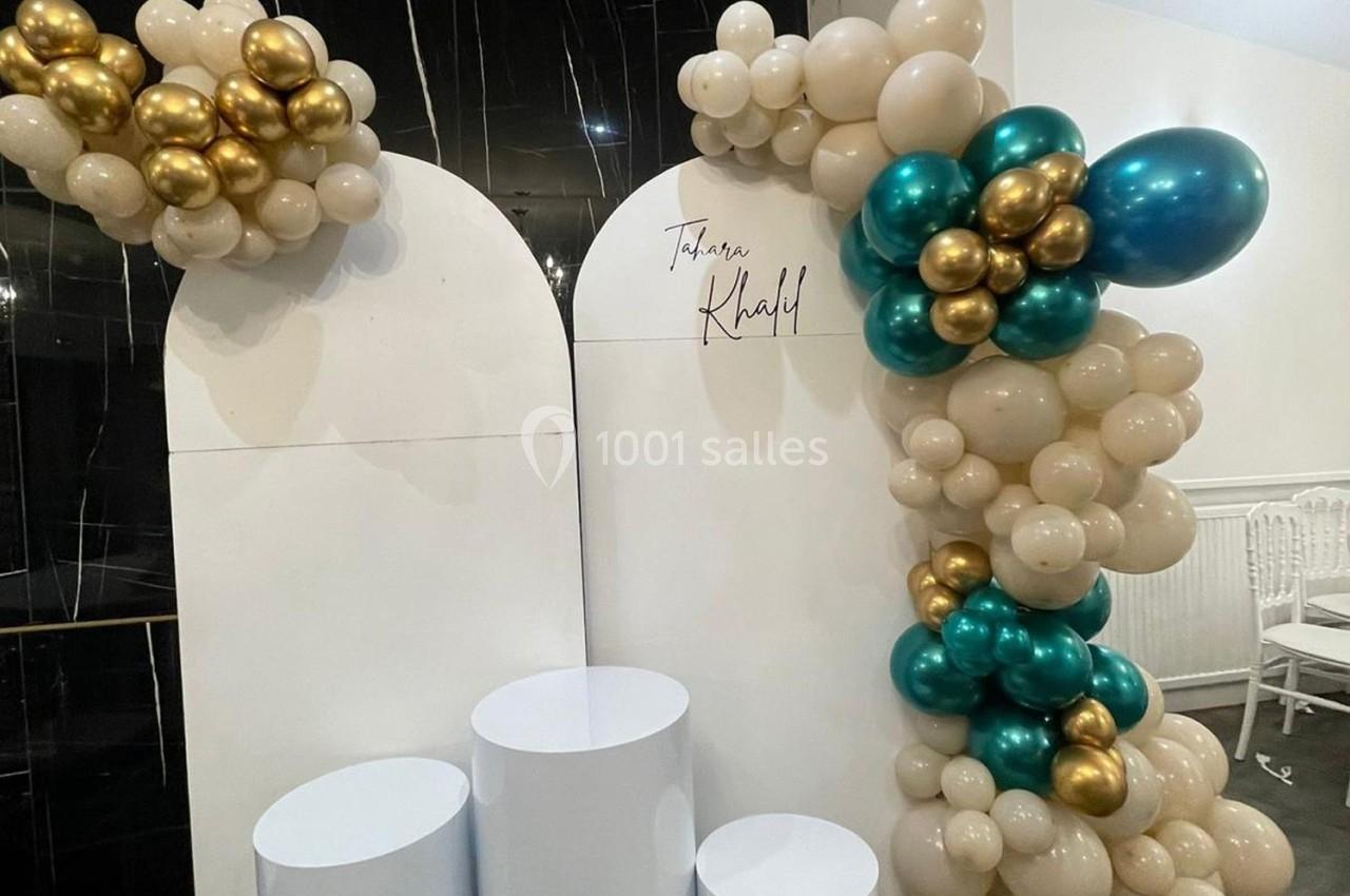 Décoration avec des ballons beiges, dorés et verts autour de panneaux blancs et colonnes cylindriques.