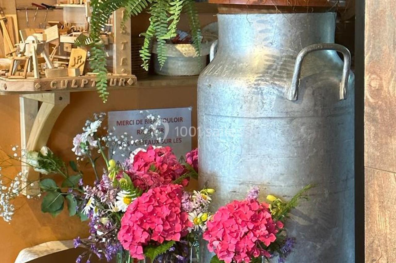 Un grand bidon en métal entouré de fleurs colorées et de plantes, avec des objets en bois en arrière-plan.