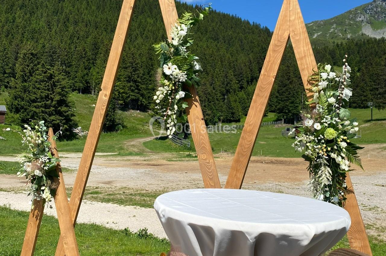 Structure en bois décorée de fleurs blanches et vertes, installée en extérieur avec vue sur des montagnes et une prairie.