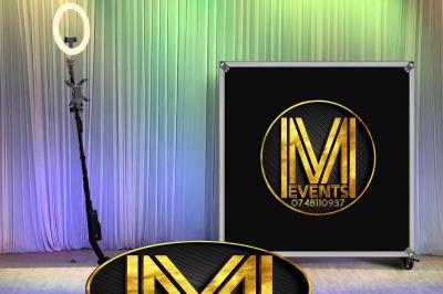 Un anneau lumineux sur trépied et un panneau noir avec un logo doré ’IM Events’ devant un rideau coloré.