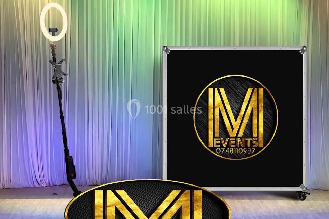 Un anneau lumineux sur trépied et un panneau noir avec un logo doré ’IM Events’ devant un rideau coloré.