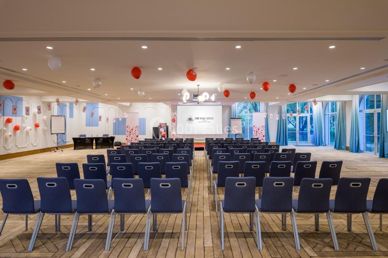 Salle de conférence avec des rangées de chaises, un écran de projection, des décorations rouges et blanches.