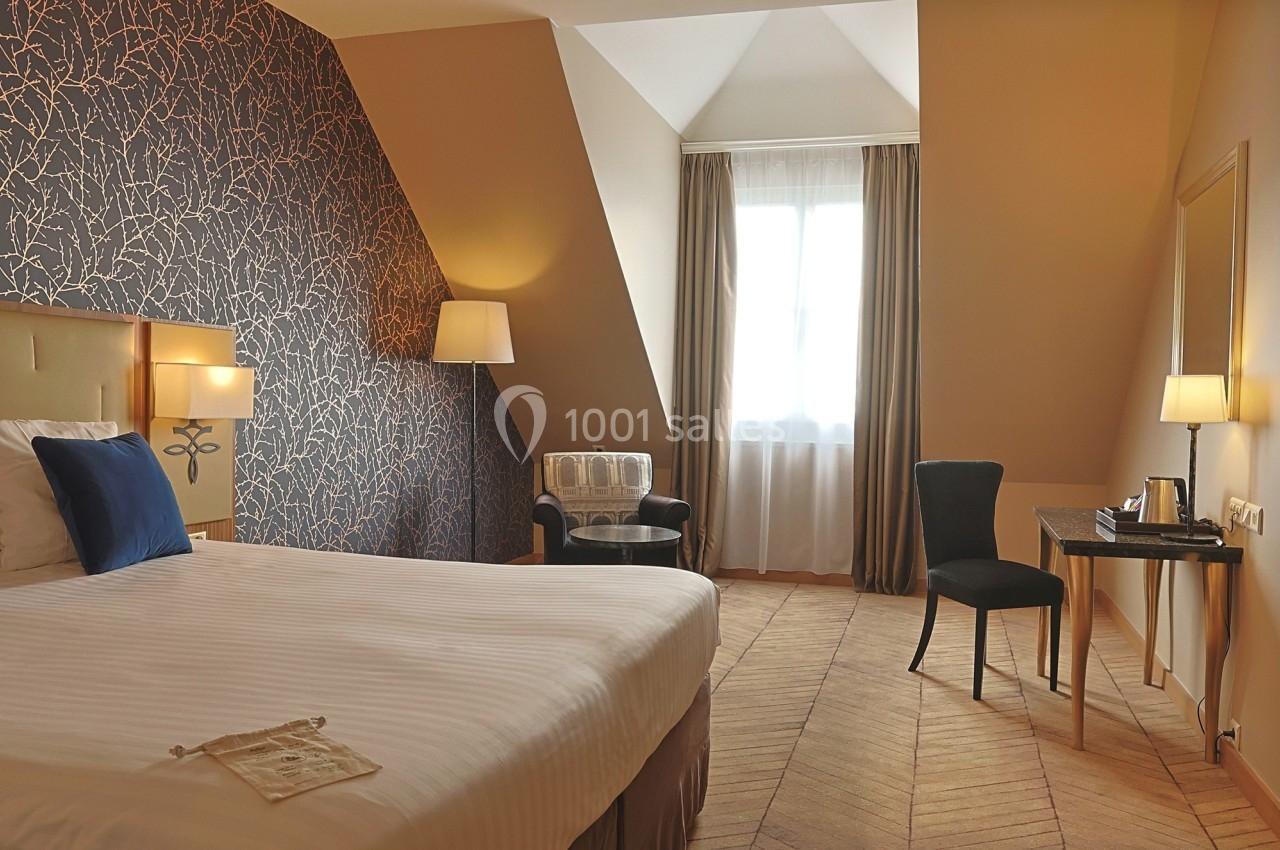 Chambre lumineuse sous combles avec lit double, bureau, fauteuil, lampes et fenêtre avec rideaux beiges.