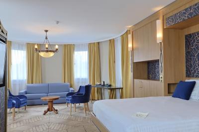 Chambre d'hôtel lumineuse avec lit double, coin salon, rideaux jaunes, mobilier en bois et fauteuils bleus.