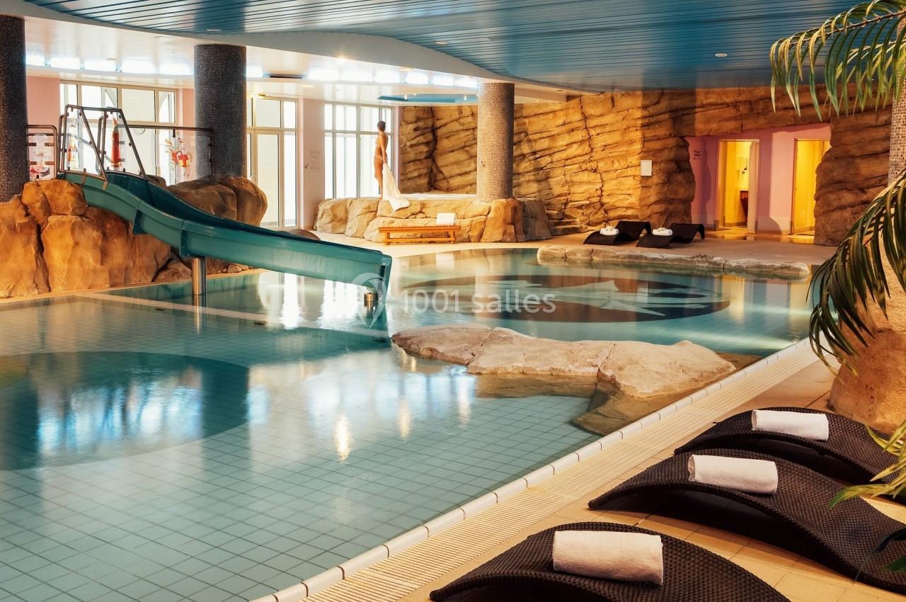Piscine intérieure avec toboggan, rochers décoratifs, transats et éclairage naturel.