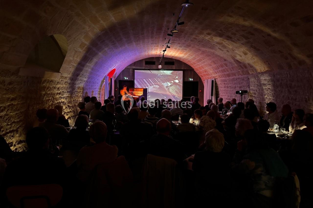Salle voûtée avec un public assis face à une scène éclairée où se déroule une performance artistique.