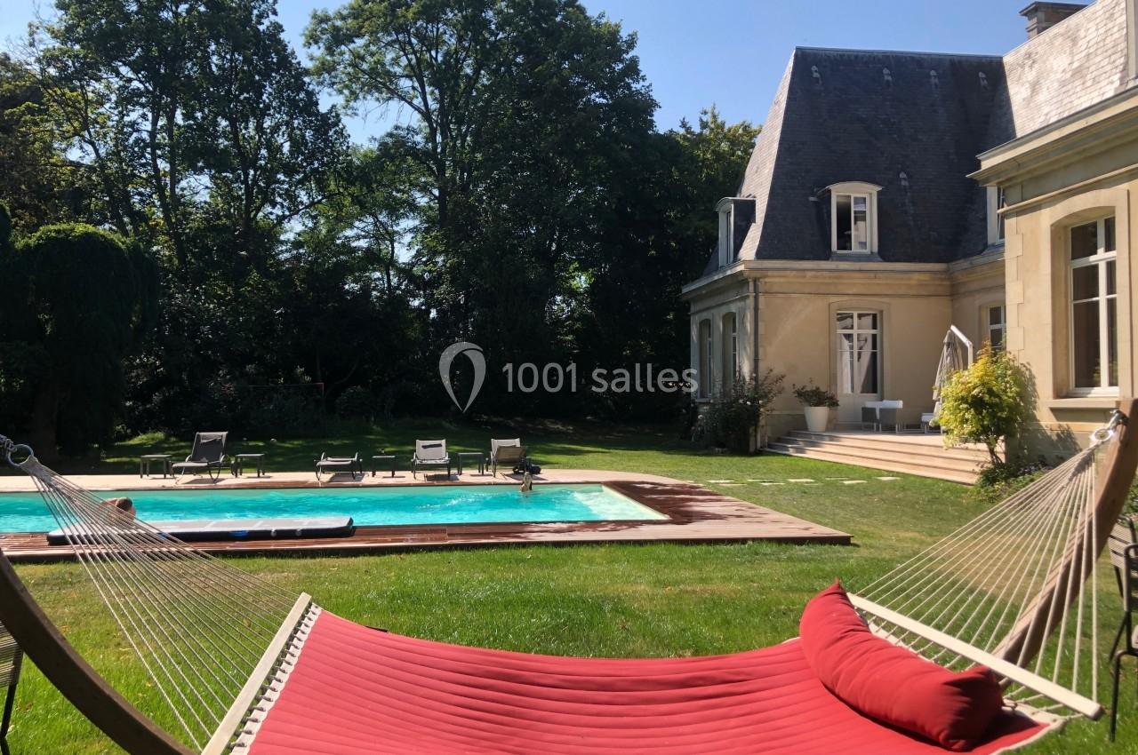 Hamac rouge installé dans un jardin avec piscine, entouré de chaises longues et d'une grande maison en arrière-plan.