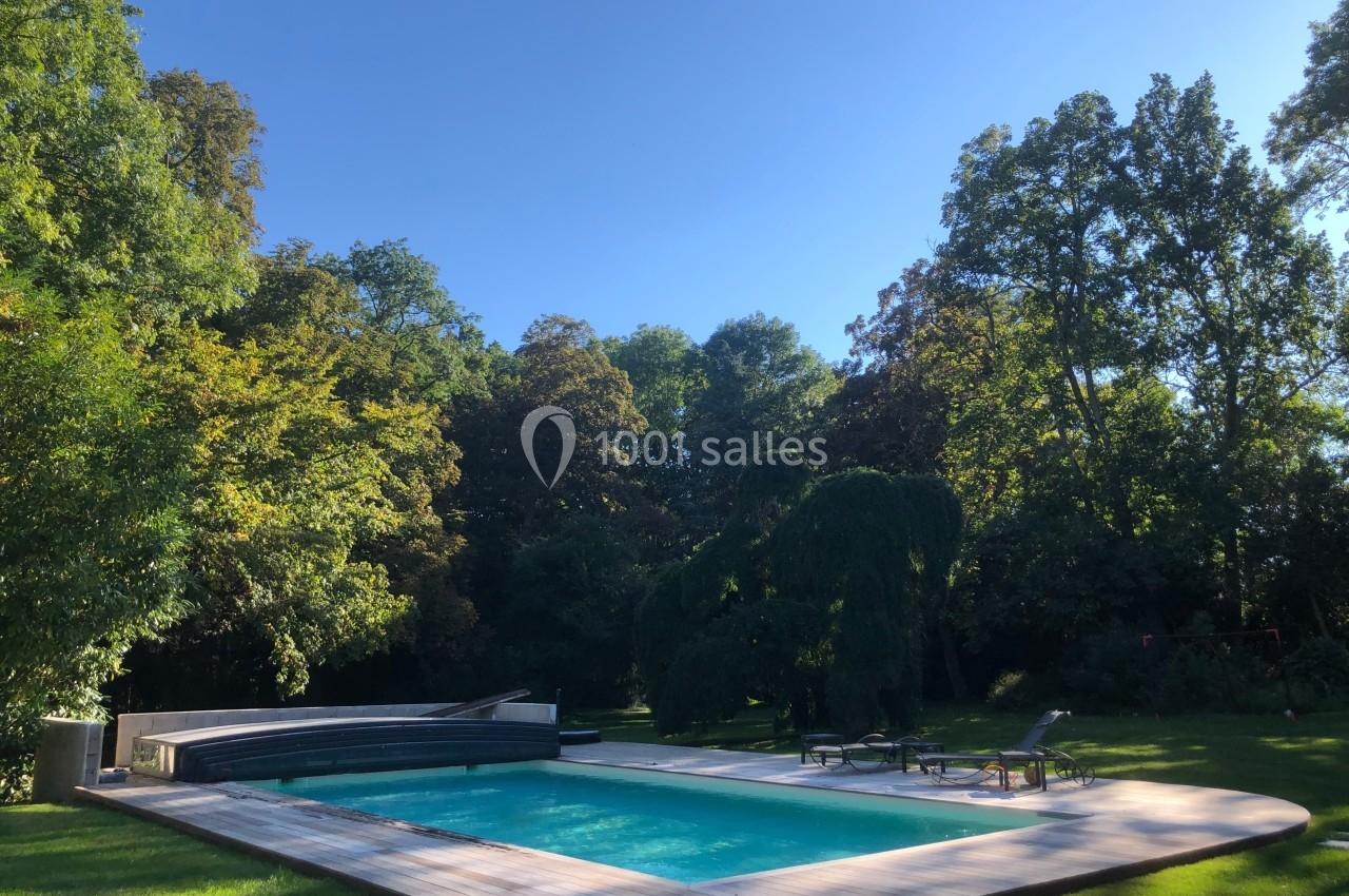 Piscine extérieure entourée de verdure et de chaises longues, sous un ciel bleu dégagé.