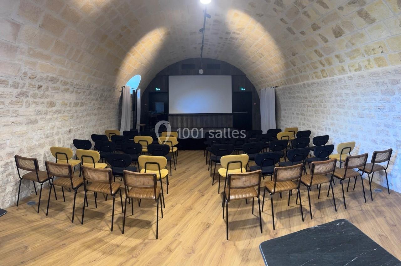 Salle voûtée en pierre avec des rangées de chaises disposées face à un écran de projection.