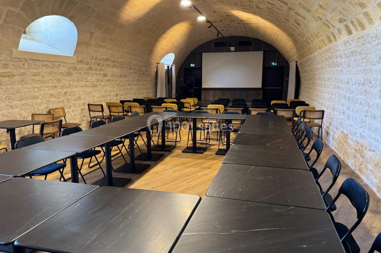 Salle de réunion voûtée avec murs en pierre, tables noires disposées en U et écran de projection au fond.