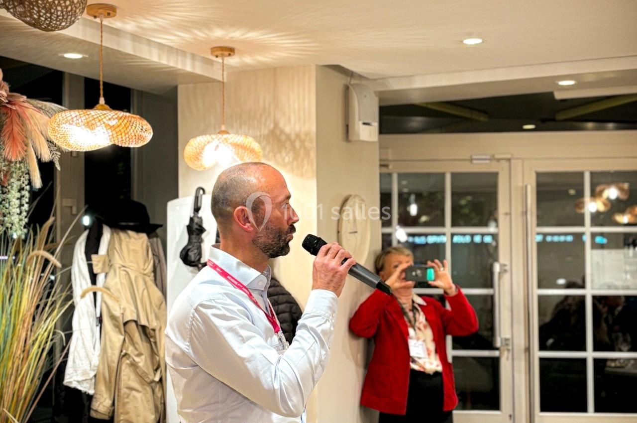 Un homme debout parle dans un micro dans une salle éclairée, tandis qu'une femme prend une photo en arrière-plan.