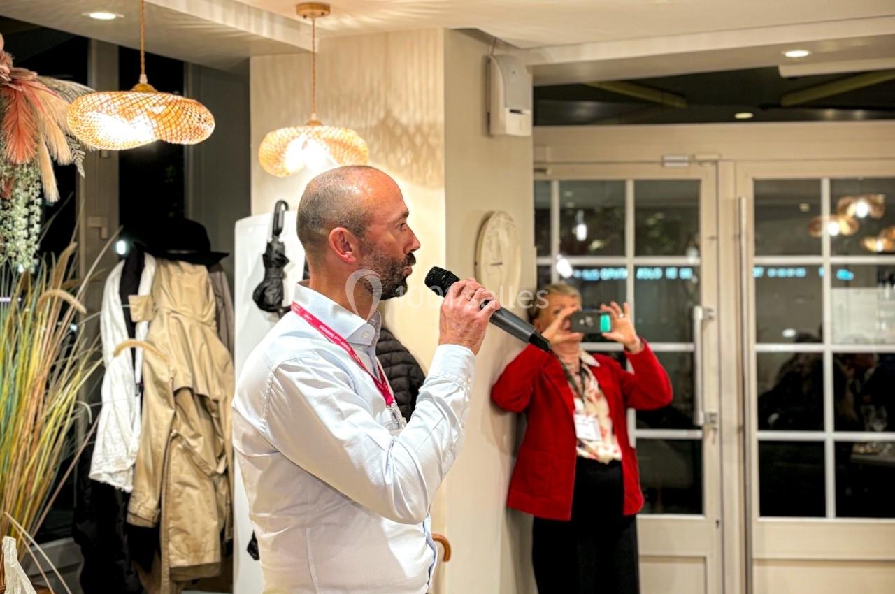 Un homme parle dans un micro dans une pièce lumineuse, tandis qu'une femme prend une photo avec son téléphone.