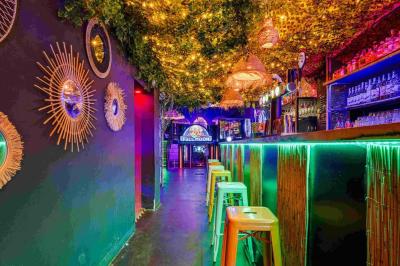 Façade d'un bar avec une décoration végétale, des néons roses et des tabourets noirs sur le trottoir.