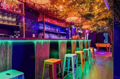 Façade d'un bar avec une décoration végétale, des néons roses et des tabourets noirs sur le trottoir.