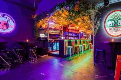 Façade d'un bar avec une décoration végétale, des néons roses et des tabourets noirs sur le trottoir.