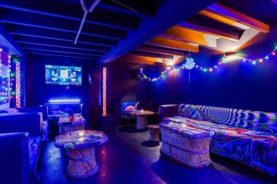 Façade d'un bar avec une décoration végétale, des néons roses et des tabourets noirs sur le trottoir.