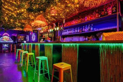 Façade d'un bar avec une décoration végétale, des néons roses et des tabourets noirs sur le trottoir.