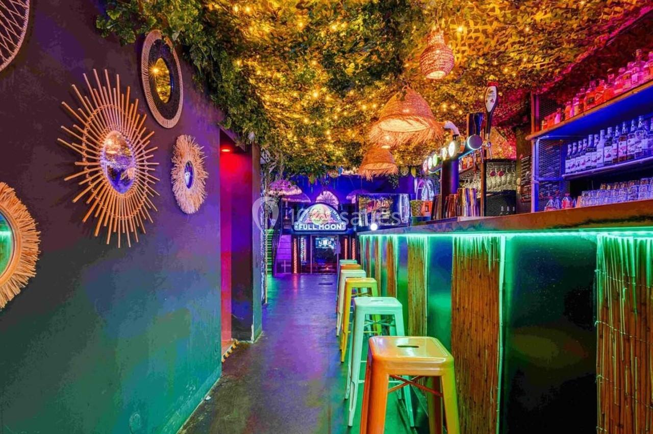 Intérieur d'un bar coloré avec un plafond végétalisé, des lumières chaleureuses et un comptoir éclairé par des néons.