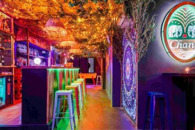 Façade d'un bar avec une décoration végétale, des néons roses et des tabourets noirs sur le trottoir.