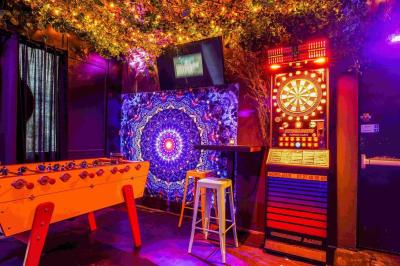 Façade d'un bar avec une décoration végétale, des néons roses et des tabourets noirs sur le trottoir.