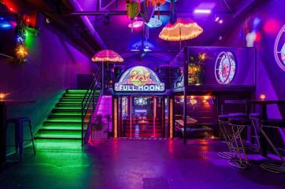 Façade d'un bar avec une décoration végétale, des néons roses et des tabourets noirs sur le trottoir.