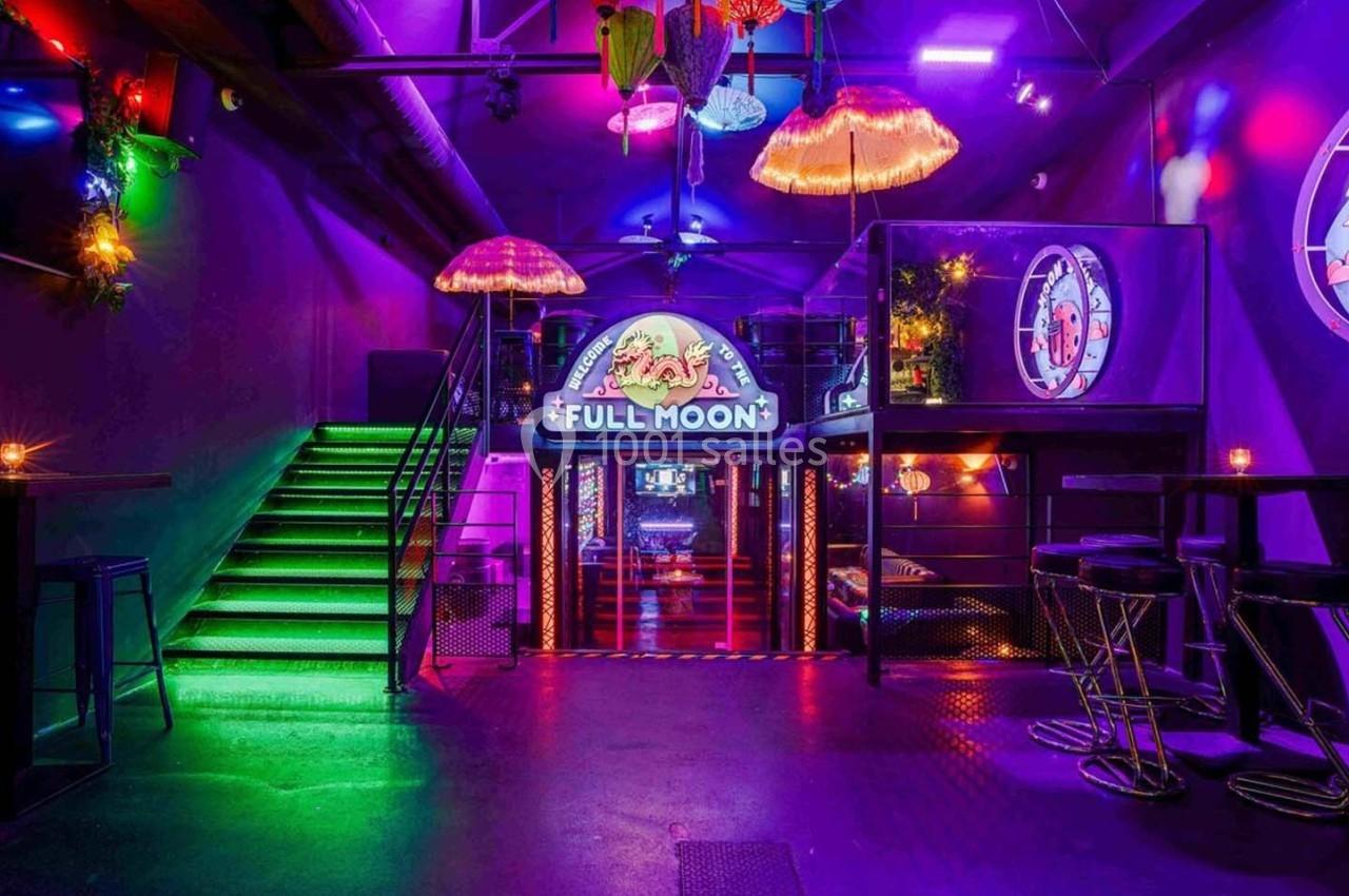 Salle de bar colorée avec éclairage néon, tables hautes, tabourets et enseigne lumineuse ’Full Moon’ au fond.