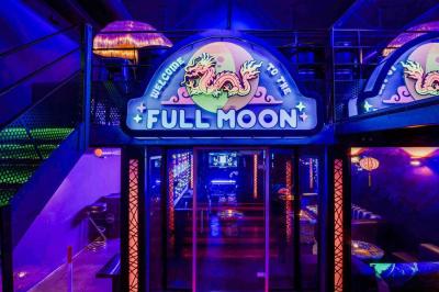 Façade d'un bar avec une décoration végétale, des néons roses et des tabourets noirs sur le trottoir.