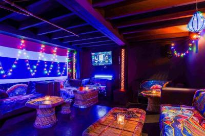 Façade d'un bar avec une décoration végétale, des néons roses et des tabourets noirs sur le trottoir.