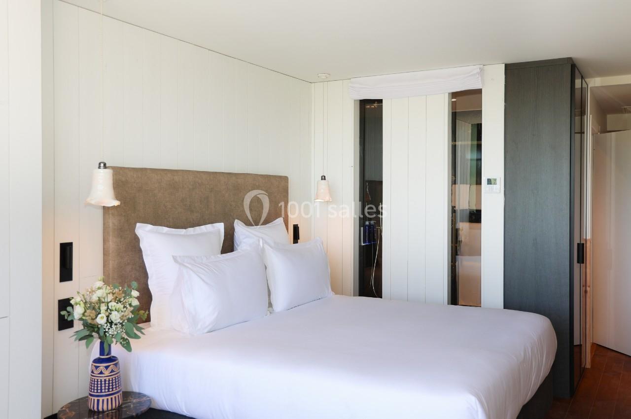 Lit double avec tête de lit rembourrée, draps blancs, lampes suspendues et vase de fleurs dans une chambre moderne.