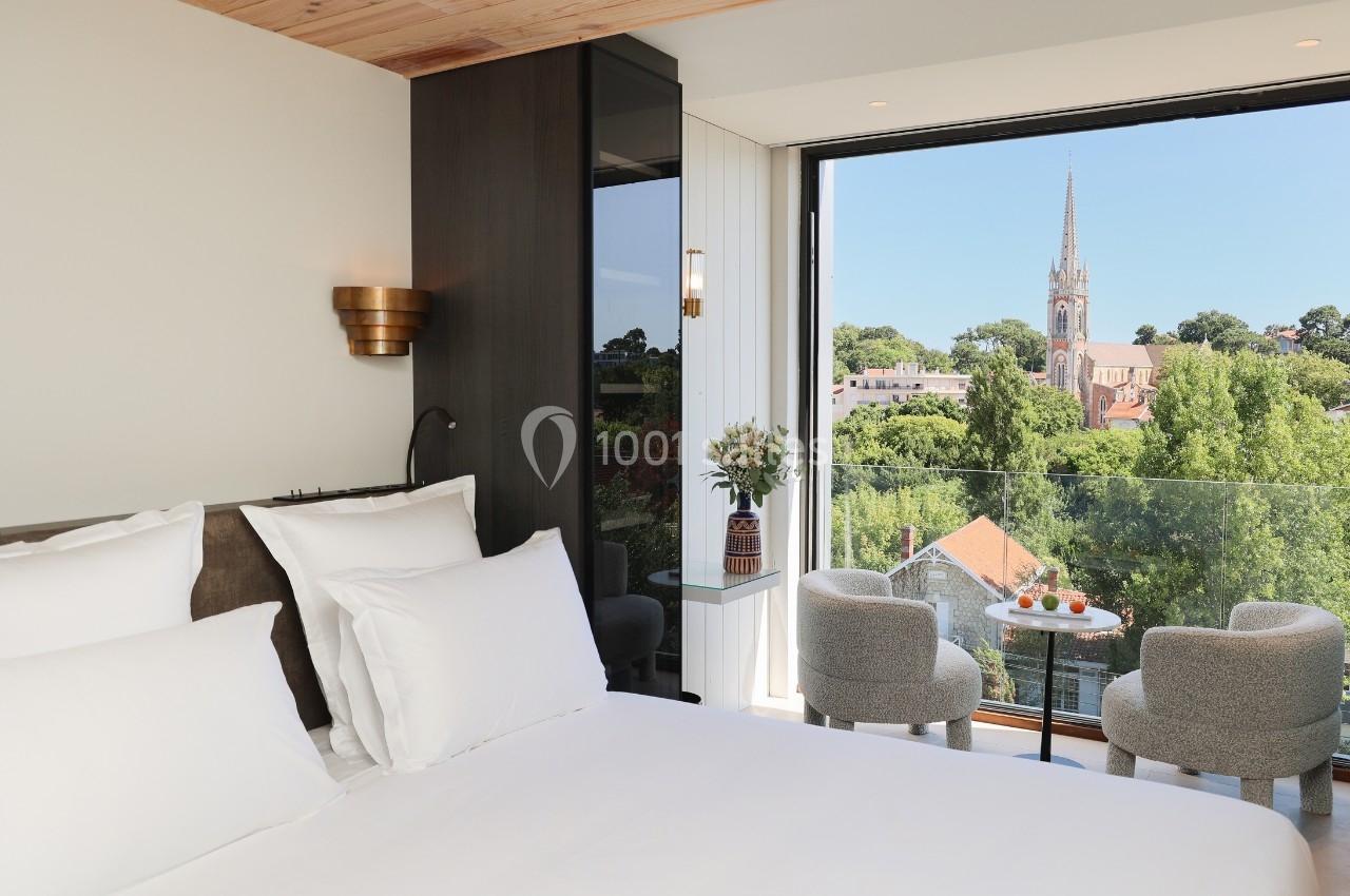 Chambre lumineuse avec lit double, balcon aménagé et vue sur un village verdoyant avec une église au loin.