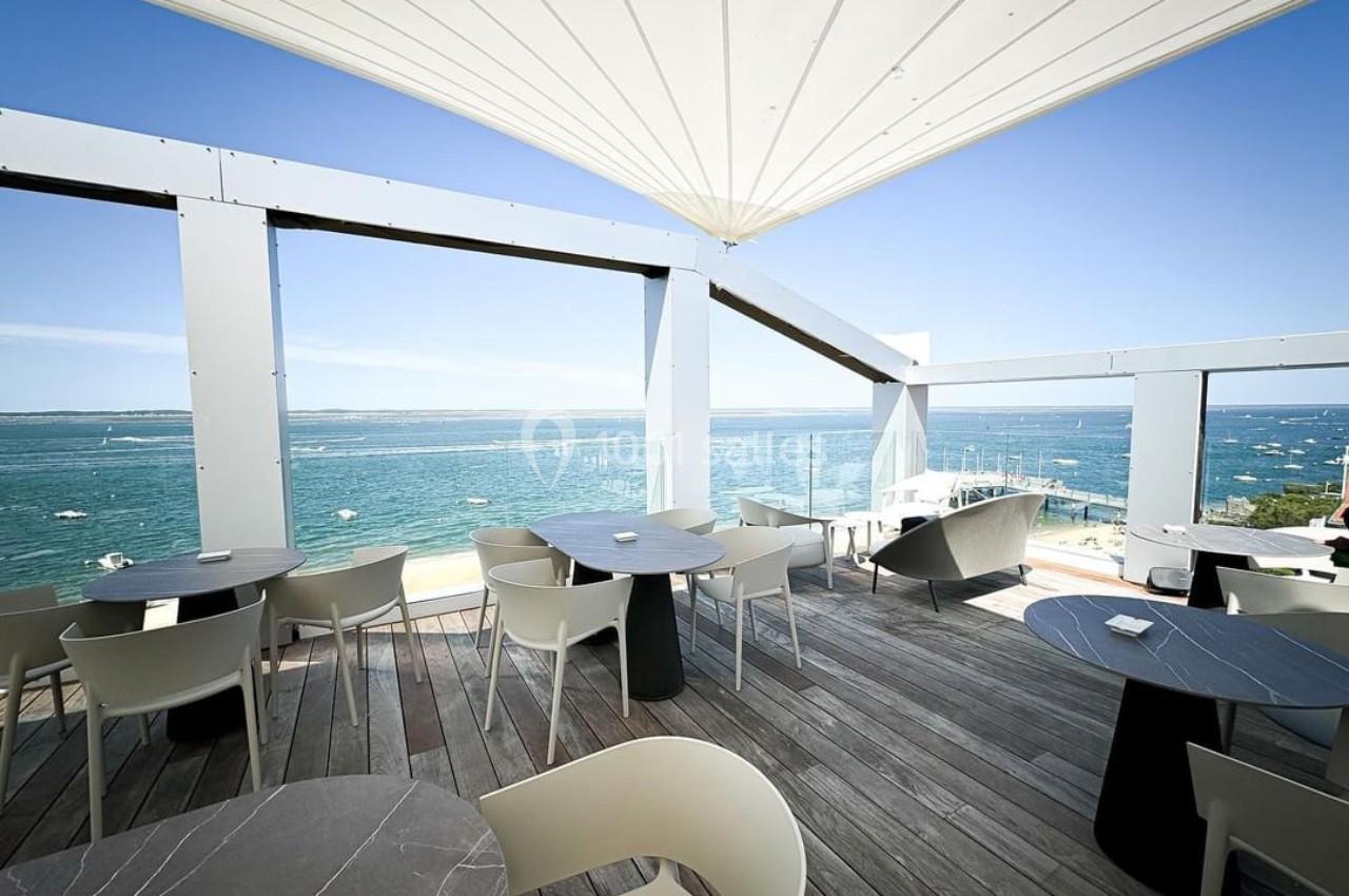 Terrasse en bois avec tables et chaises, offrant une vue dégagée sur la mer et un ciel bleu ensoleillé.