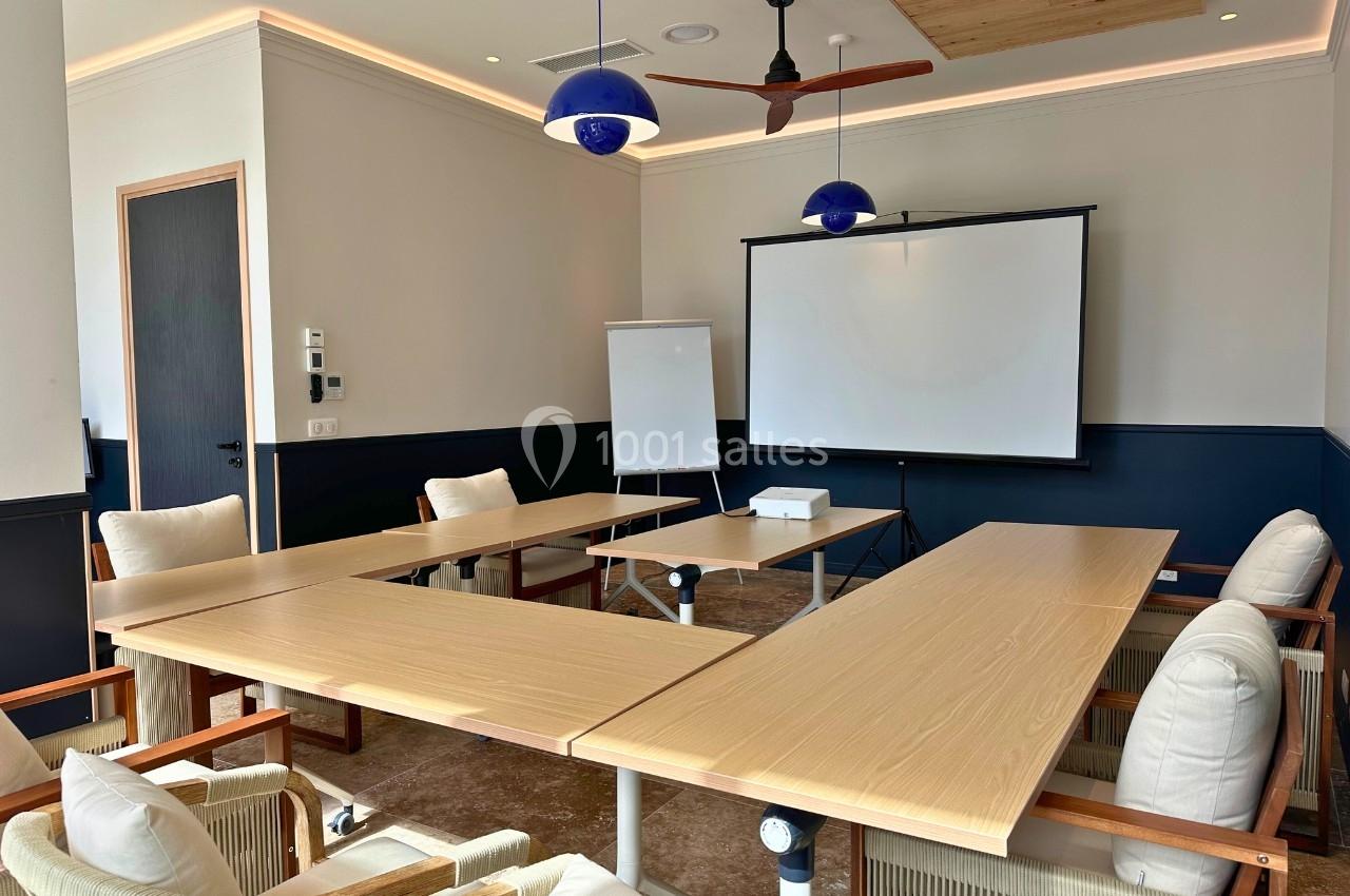 Salle de réunion lumineuse avec tables en bois disposées en U, chaises rembourrées, tableau blanc et écran de projection.