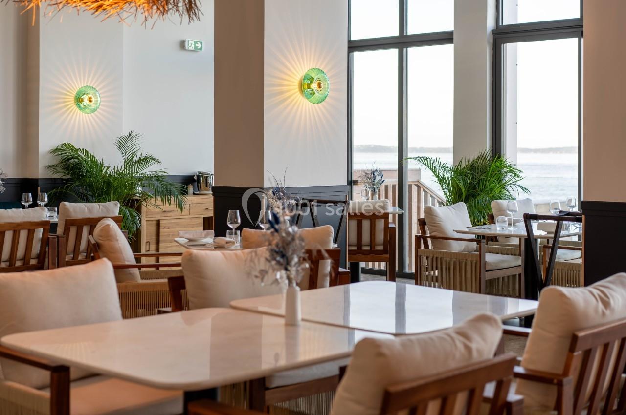 Salle de restaurant lumineuse avec tables en marbre, chaises en bois, plantes vertes et vue sur l'extérieur.