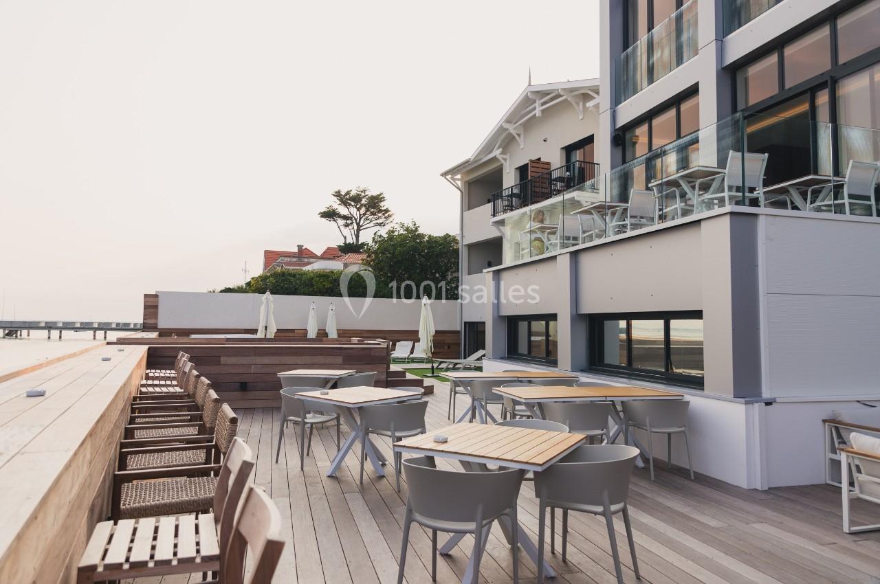 Terrasse extérieure avec tables, chaises et parasols, donnant sur un bâtiment moderne et un paysage côtier.