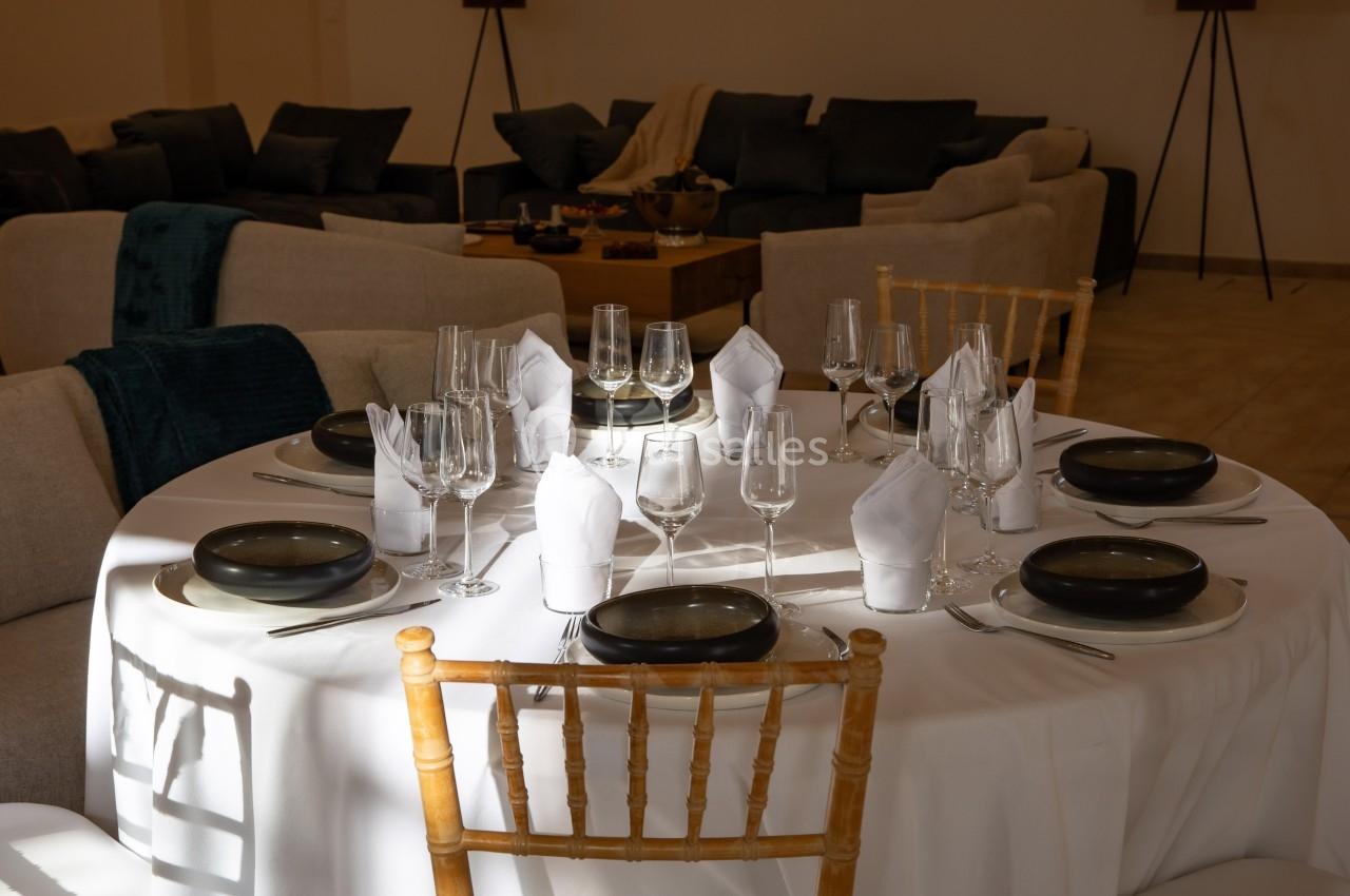Table ronde dressée avec une nappe blanche, vaisselle noire, verres à pied et serviettes pliées, entourée de chaises.