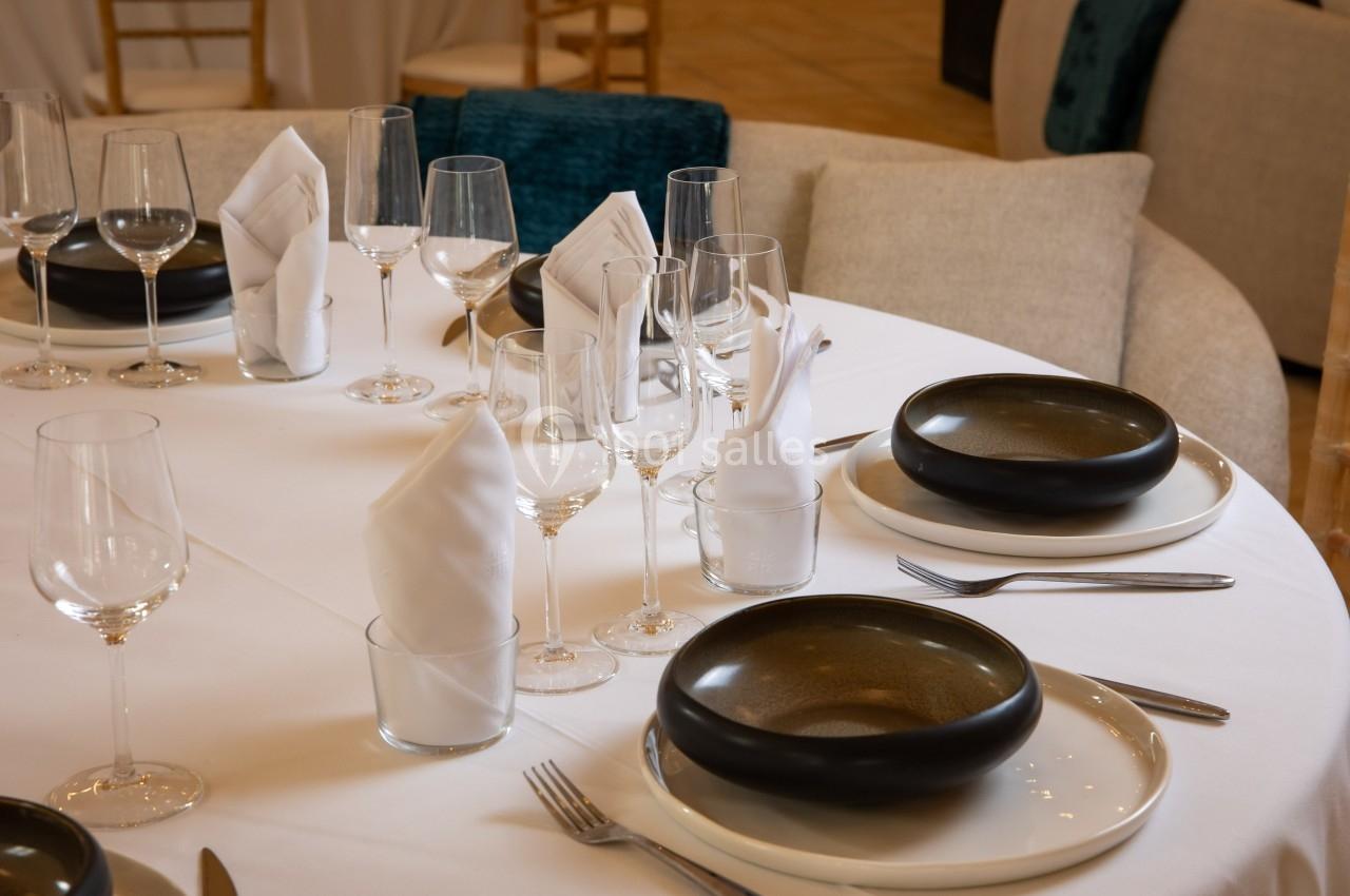 Table dressée avec nappes blanches, assiettes noires, verres à pied et serviettes pliées dans un cadre élégant.