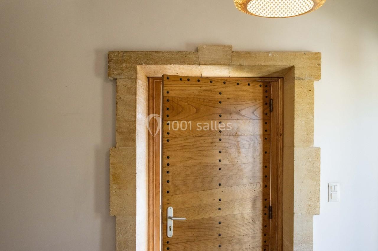 Porte en bois massif légèrement entrouverte, encadrée de pierre, sous une suspension en rotin dans une pièce lumineuse.