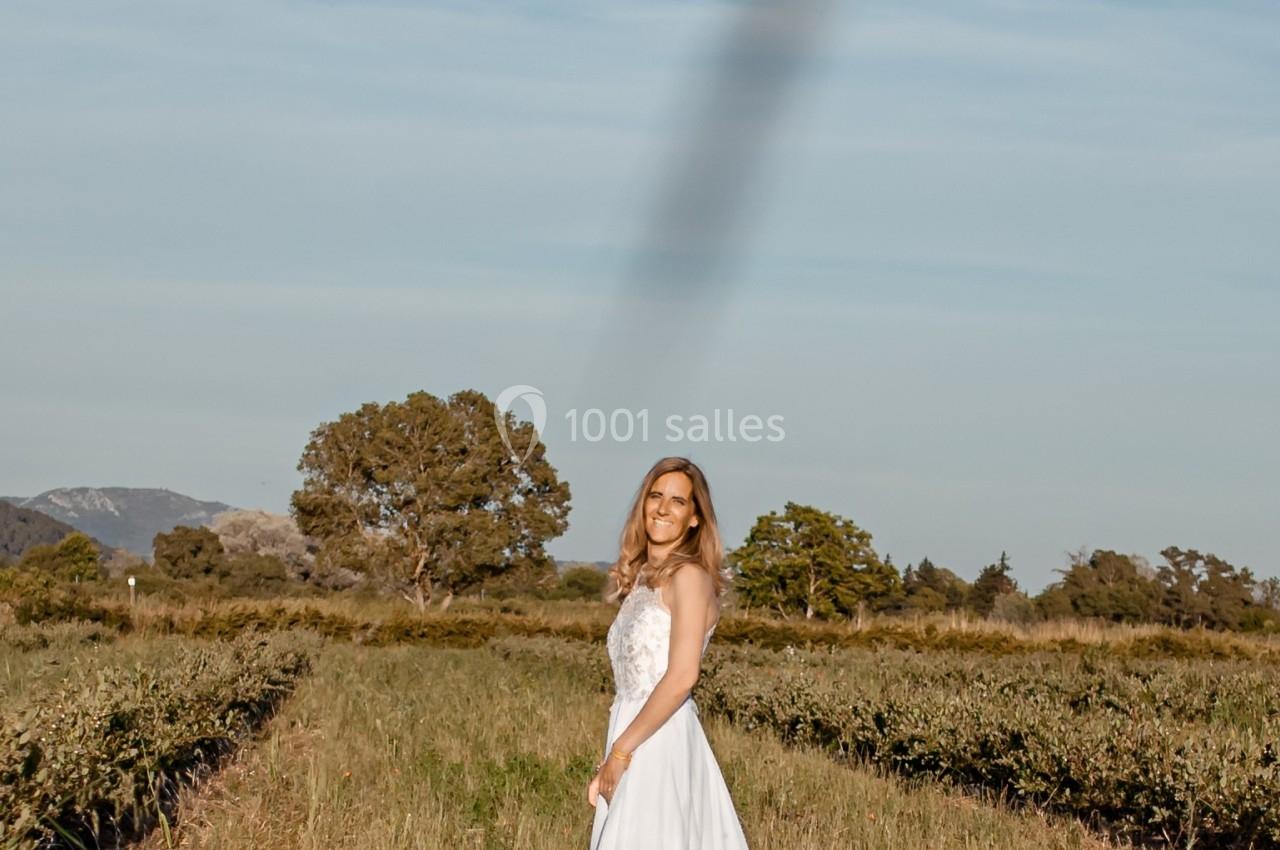 Femme en robe blanche debout dans un champ, entourée de végétation sous un ciel dégagé.